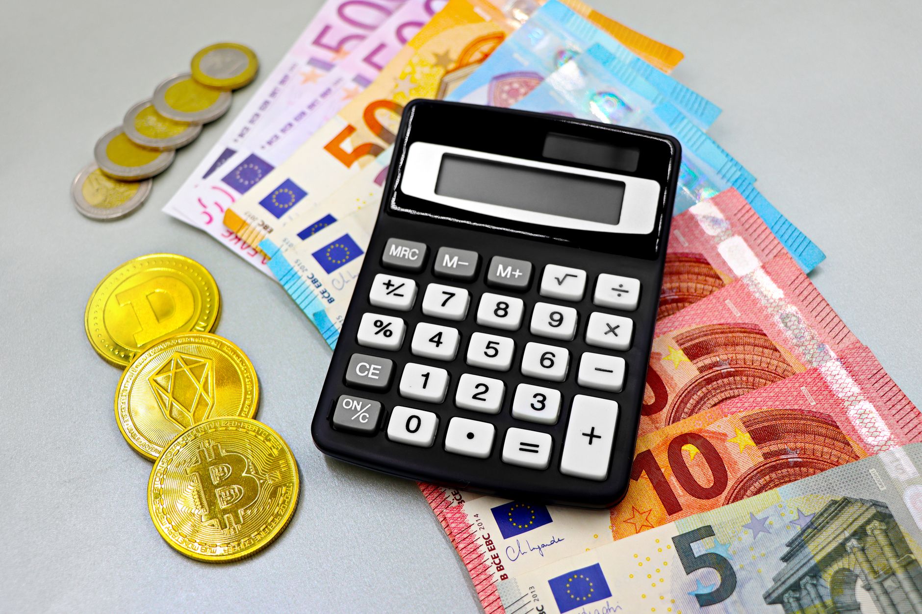 Calculatrice noire posée sur des billets en euros, avec des pièces et des jetons dorés de cryptomonnaies sur une table grise.