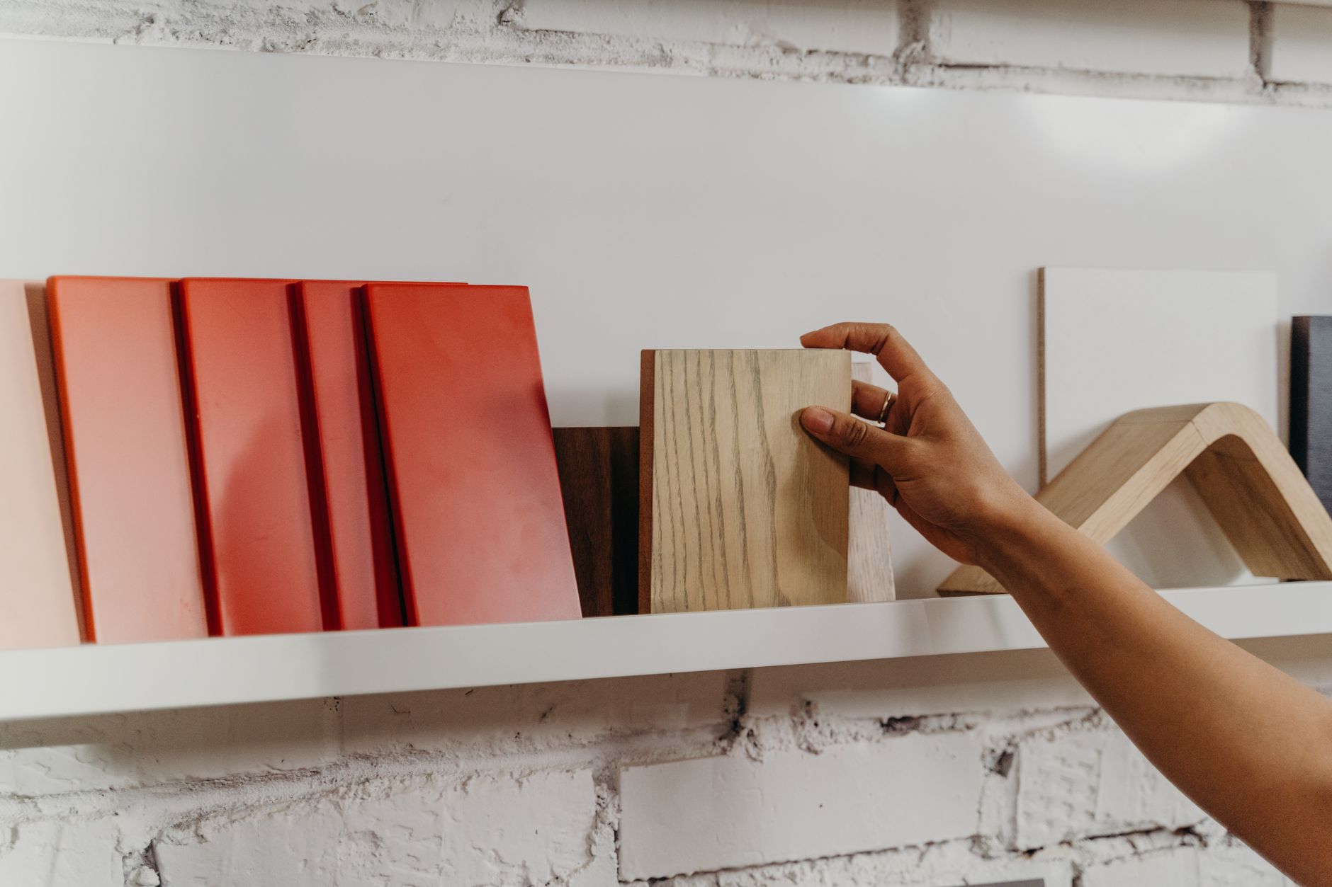 Une main attrape un échantillon de bois clair sur une étagère blanche, près de plaques rouges, devant un mur de briques blanches.