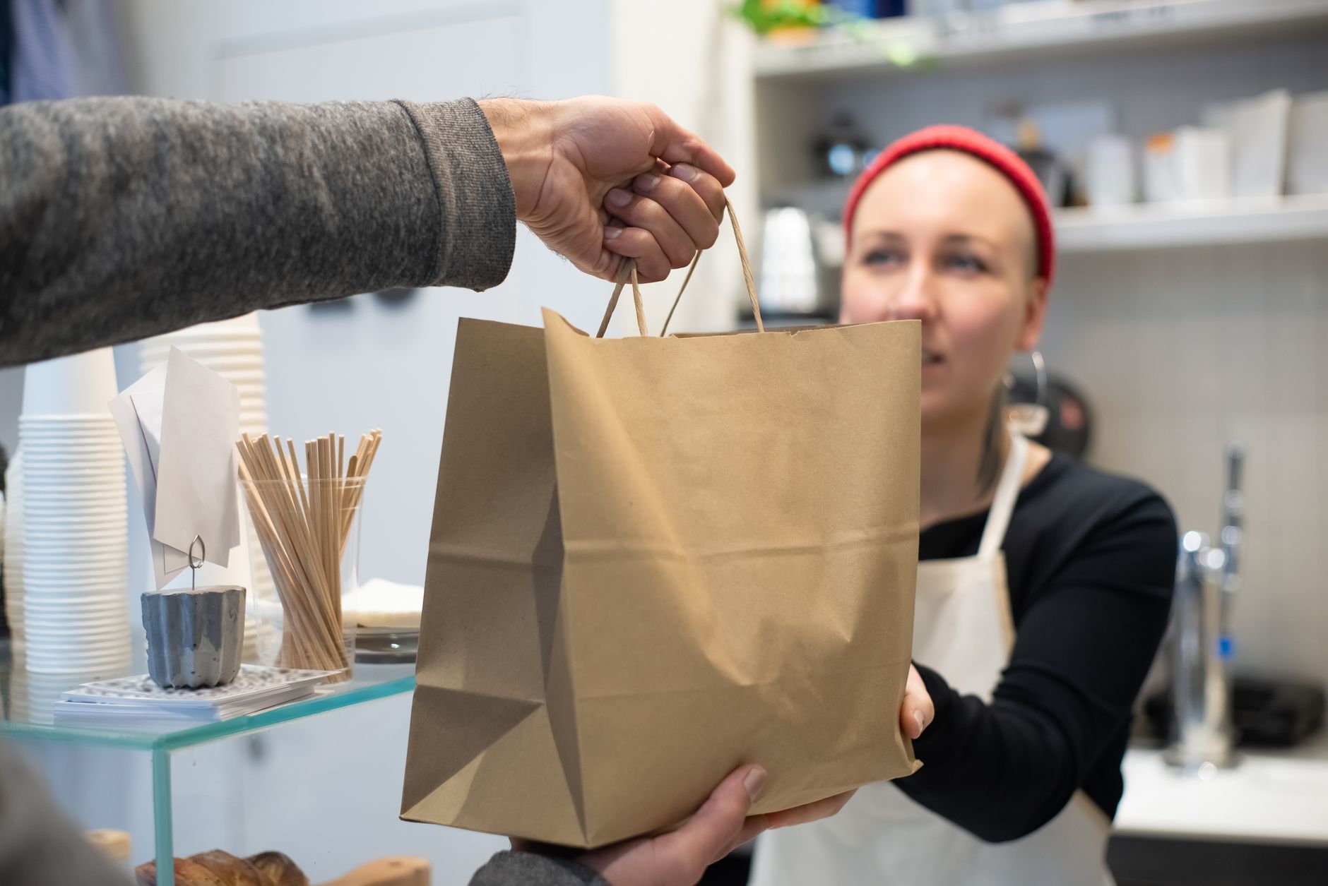 Une vendeuse en tablier tend un sac en papier kraft à un client au comptoir d’un petit commerce de restauration.